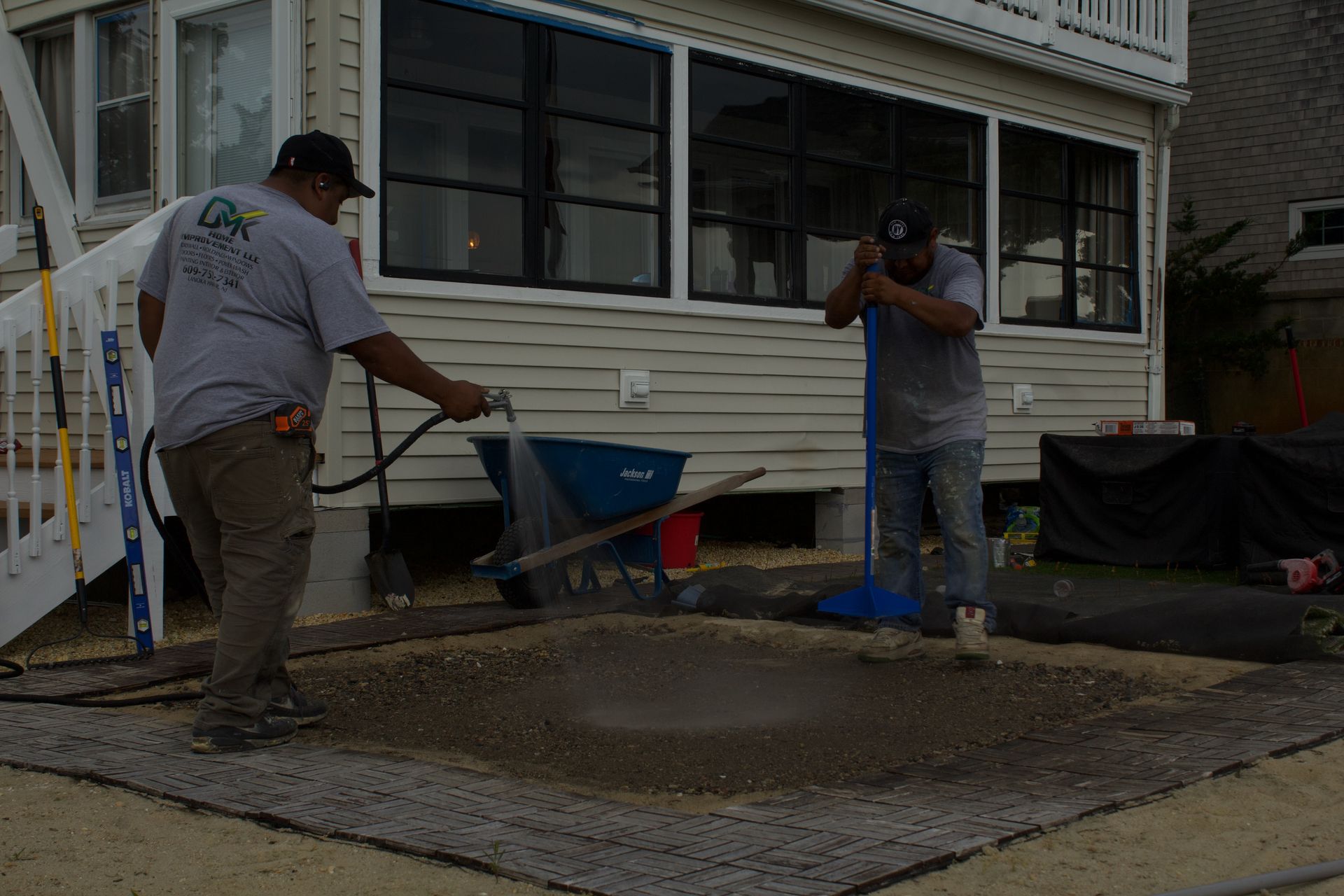 Two workers pouring and leveling concrete beside a house, with wheelbarrows and tools nearby.