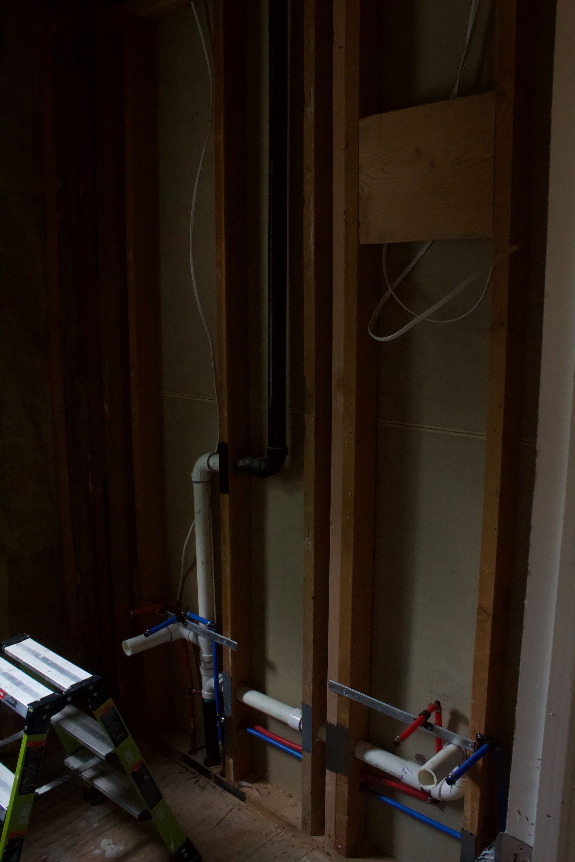 Exposed plumbing pipes and valves in an unfinished interior wall, with a ladder at left.