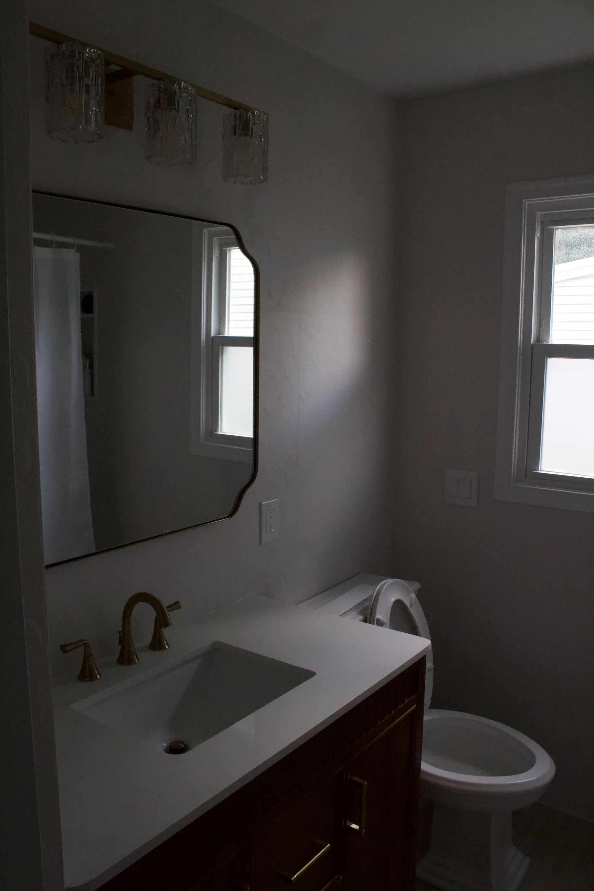 Small bathroom with white sink, mirror, toilet, and window in dim light
