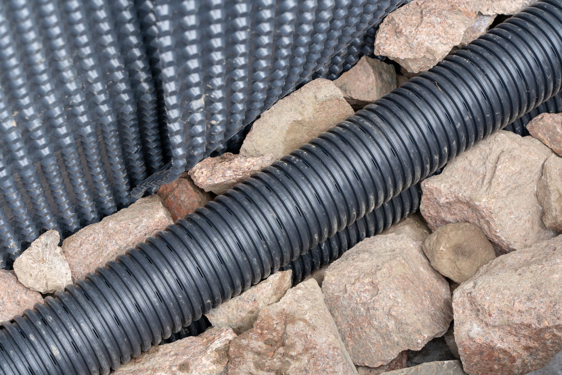 Tuyau de drainage noir ondulé entouré de pierres, à côté d'une membrane de drainage noire texturée.