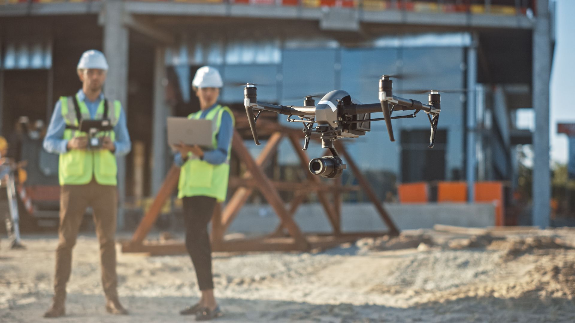 Deux ouvriers du bâtiment, vêtus de gilets de sécurité, se tiennent sur un chantier tout en pilotant un drone.