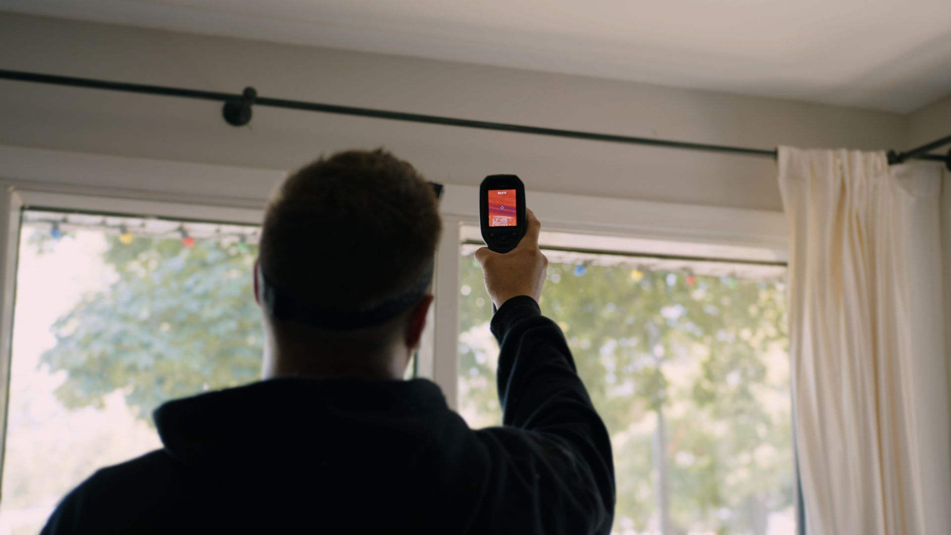 Une personne tient son téléphone devant une fenêtre, probablement en train de scanner. Intérieur lumineux avec un rideau blanc.