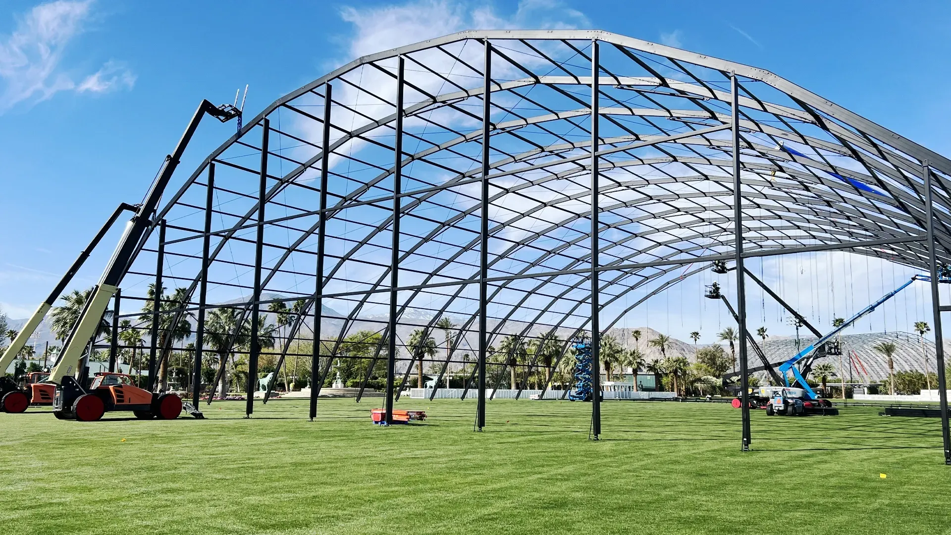 Boom lifts positioned around a large steel event structure frame with green grass and palm trees in