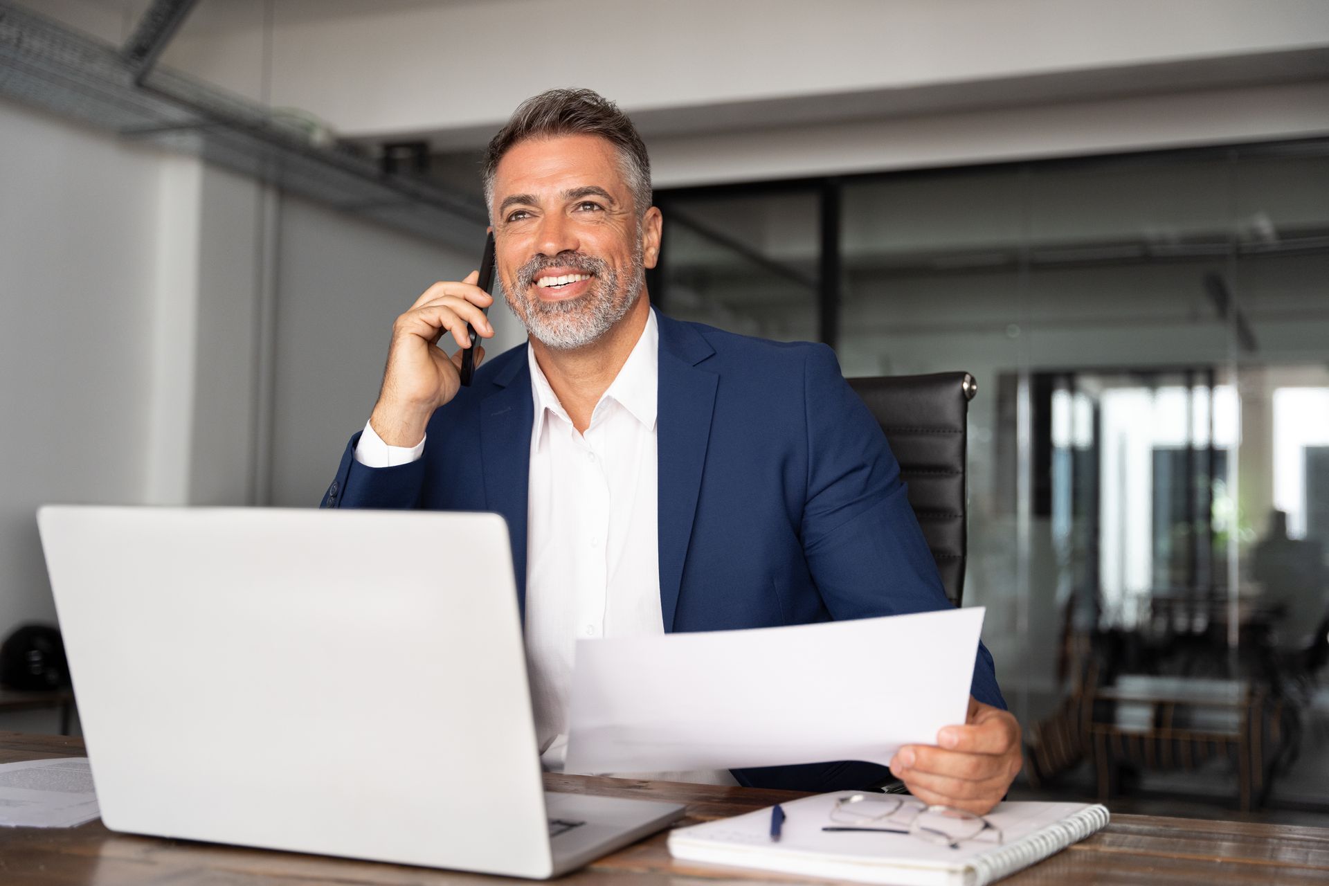 A man is sitting at a desk talking on a cell phone while holding a piece of paper.