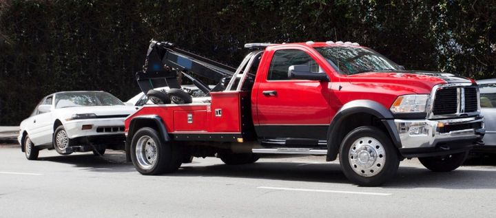 Red tow truck hauling a white car on a road, with the truck’s hood open.