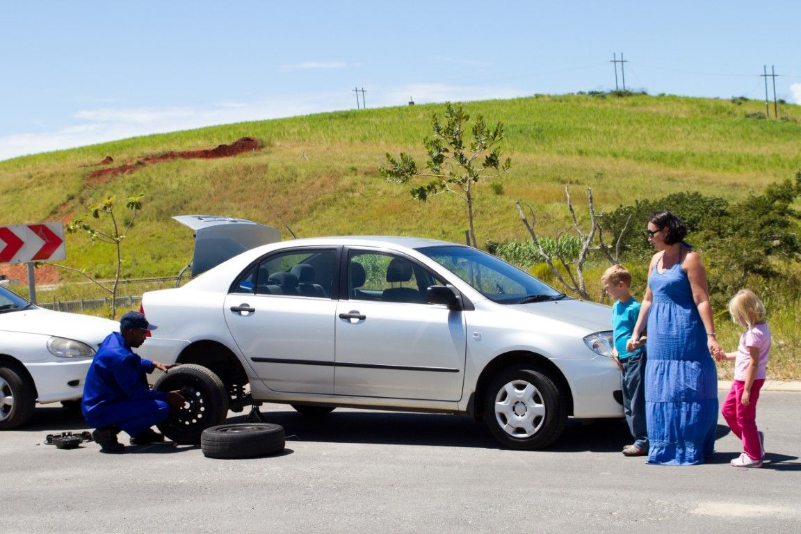 Silver car beside a road with people standing nearby in front of a grassy hill.