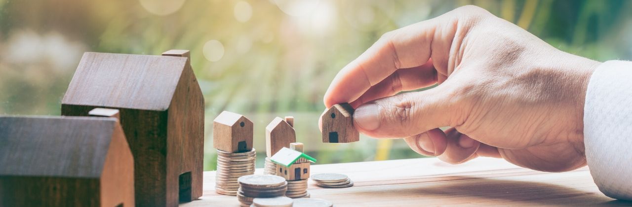 Hand placing a miniature wooden house next to others, suggesting real estate or investment.