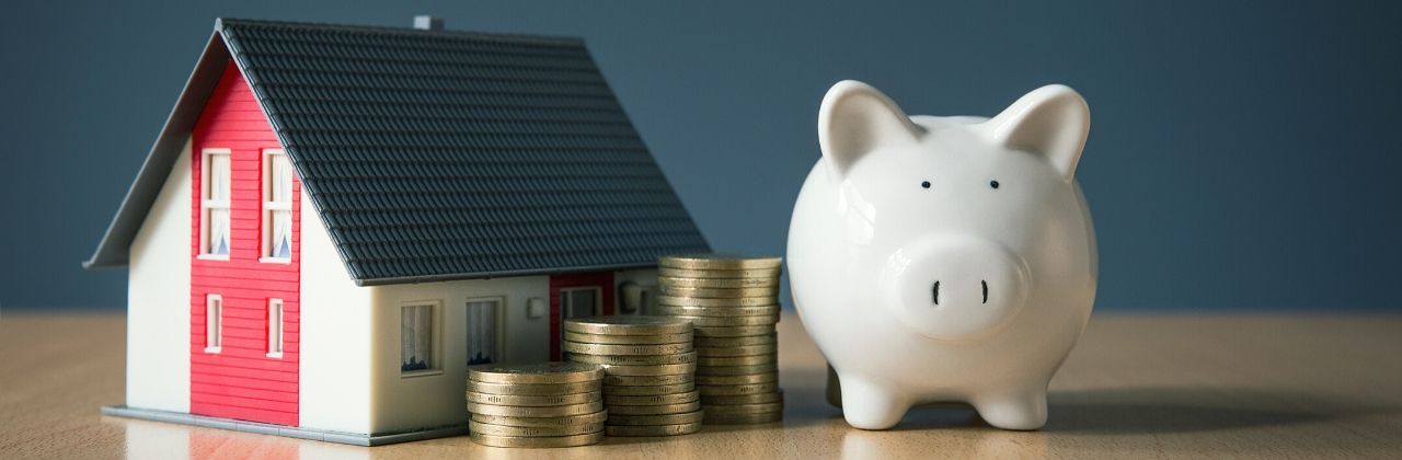 Model house, stacks of gold coins, and a white piggy bank on a wooden surface.