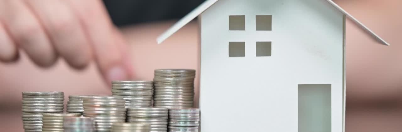 A small white paper house next to neatly stacked coins, with a blurred hand in the background.
