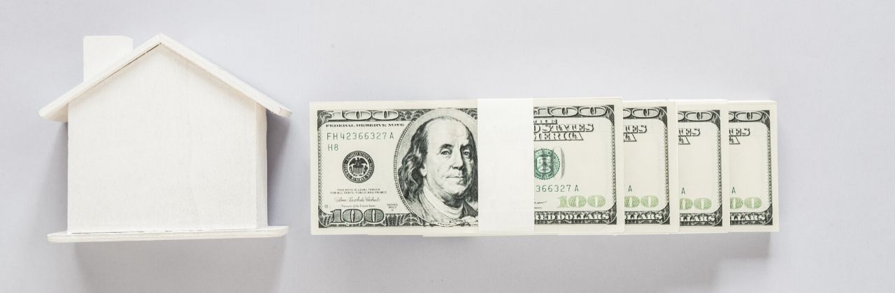 A white model house next to a stack of American hundred dollar bills.