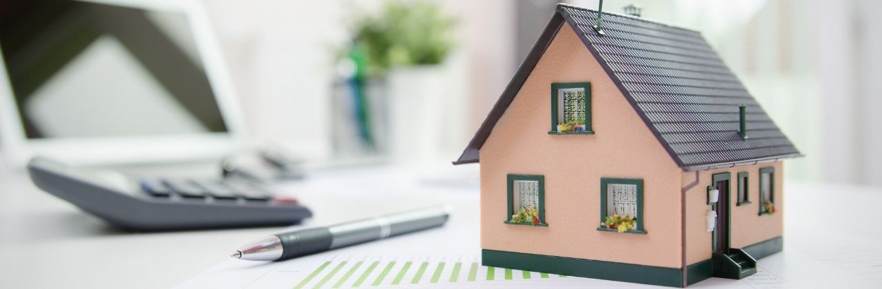 Miniature house, pen, calculator on a desk, suggesting home ownership and real estate planning.