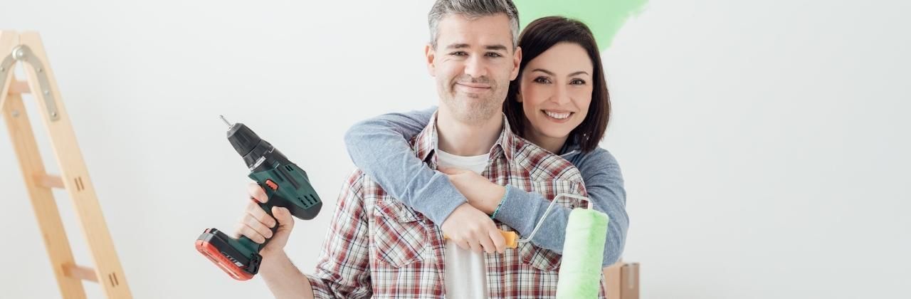 Smiling couple ready for a home renovation, holding a drill and paint roller.