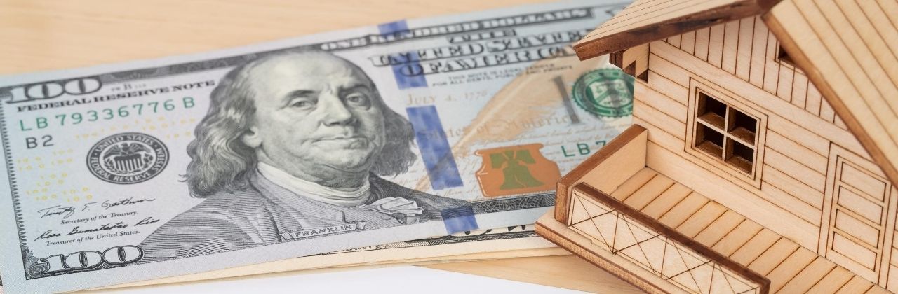 A miniature wooden house next to a hundred-dollar bill, suggesting real estate or home ownership.
