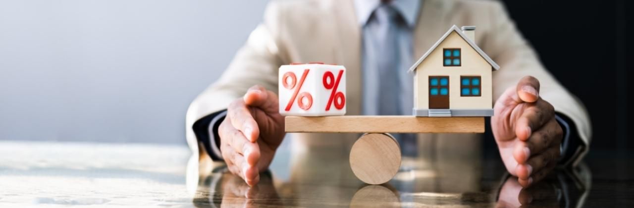 A man in a suit balances a wooden scale with a percentage block on one side and a house model on the other.