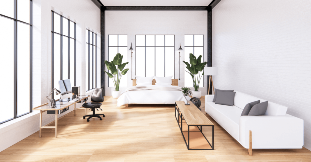 A modern, sunlit studio apartment with a bed, a desk, and a white sofa, featuring floor-to-ceiling windows and light floors.