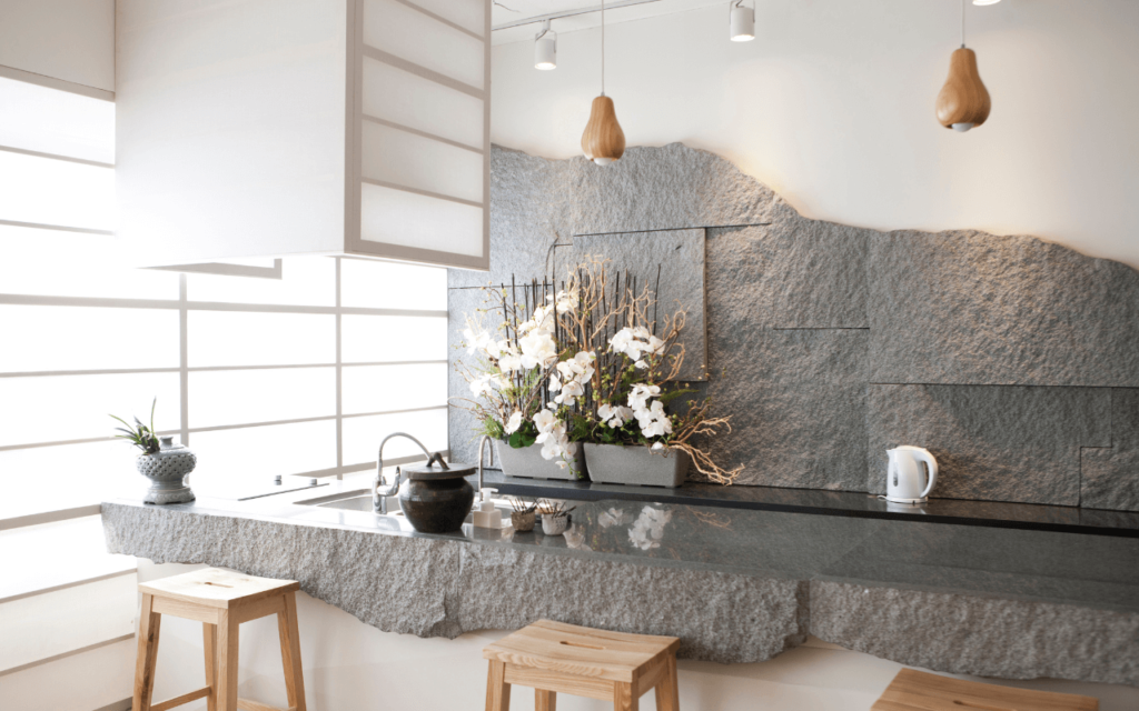 A stone kitchen counter with rough-hewn edges, a floral arrangement, wooden stools, and shoji-style window screens.