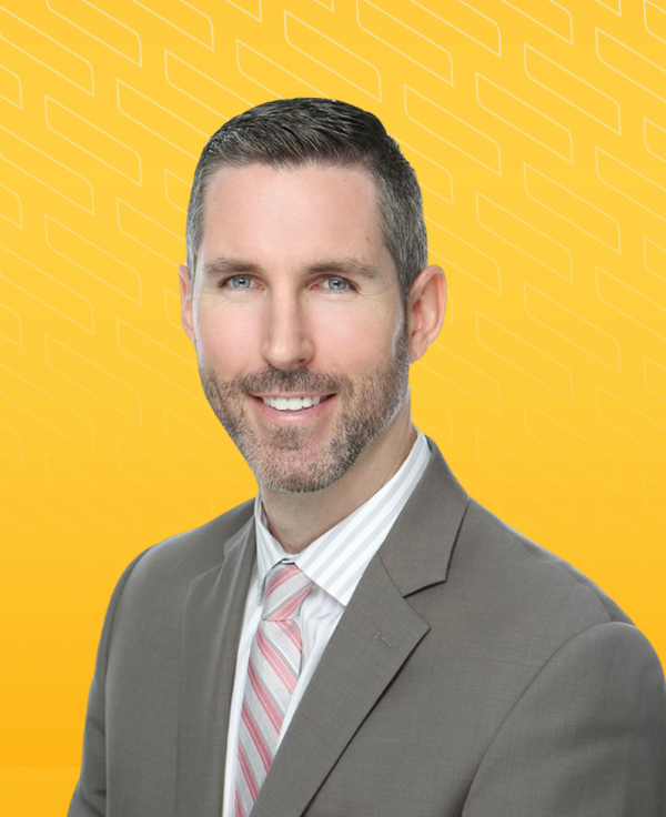 Headshot of Phil Rasmussen in a gray suit with a pink tie and pocket square, smiling against a bright yellow background.