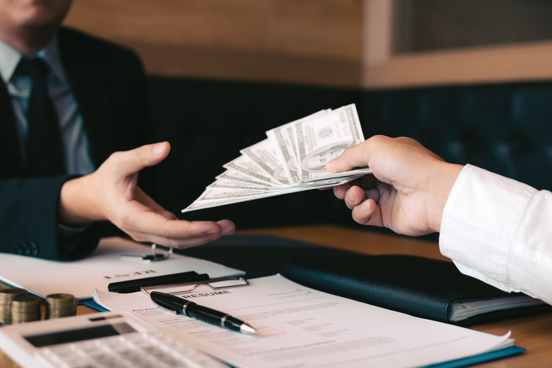 Person in suit receiving money from another person. Bills are exchanged over a desk with documents and calculator.