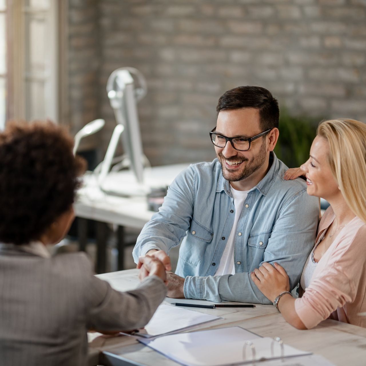 A smiling man with glasses talks with two colleagues in a modern office with a brick wall background.