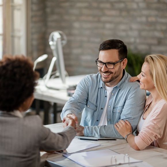 A smiling man with glasses talks with two colleagues in a modern office with a brick wall background.