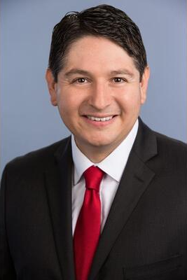 Michael Sandoval smiling in a black suit and red tie against a blue studio background.