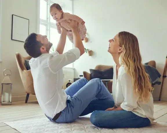 Father playfully tossing baby up in air as mother watches, all smiling inside the house.