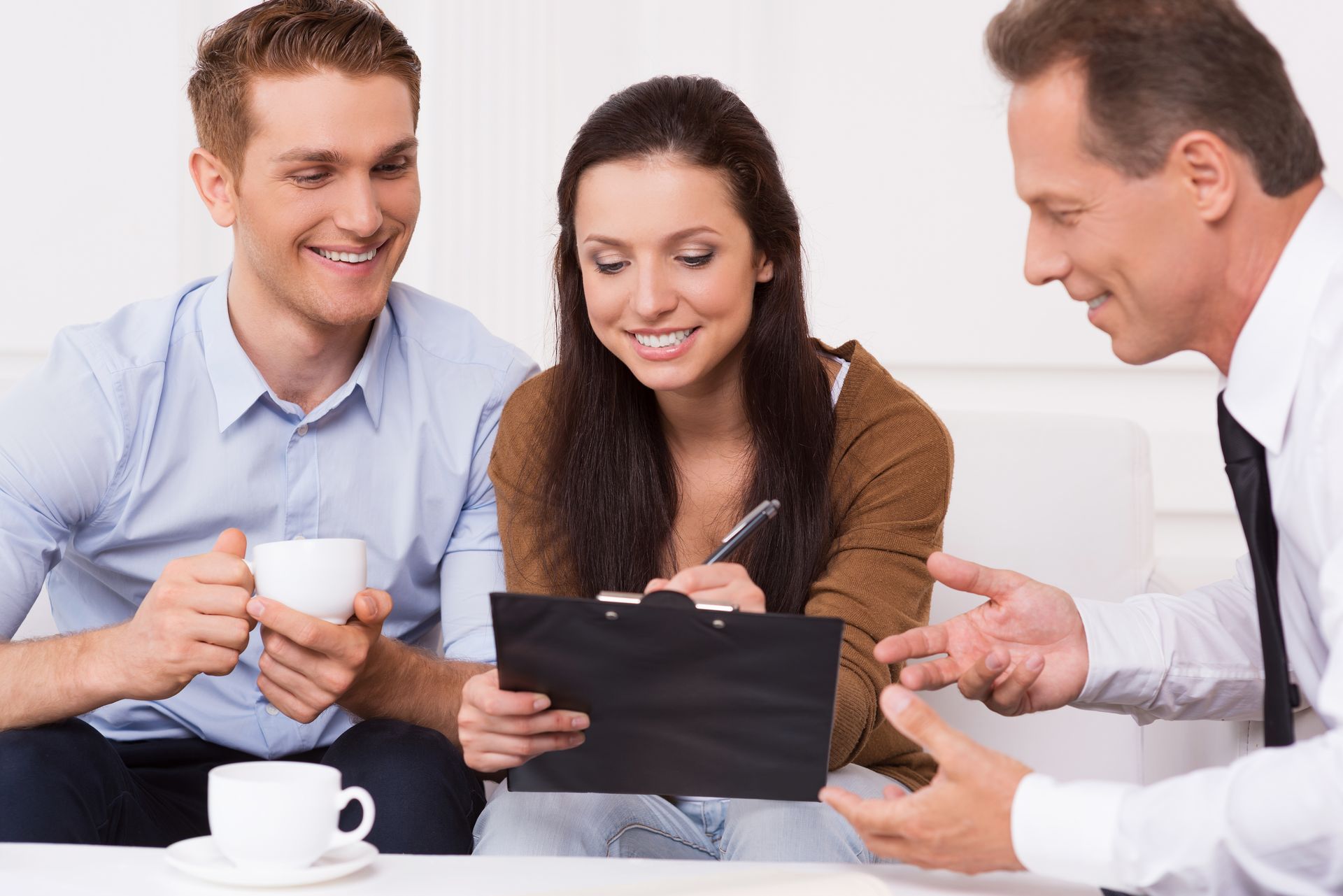 Couple signing document with advisor. Woman smiles while writing, man holds cup, advisor gestures.
