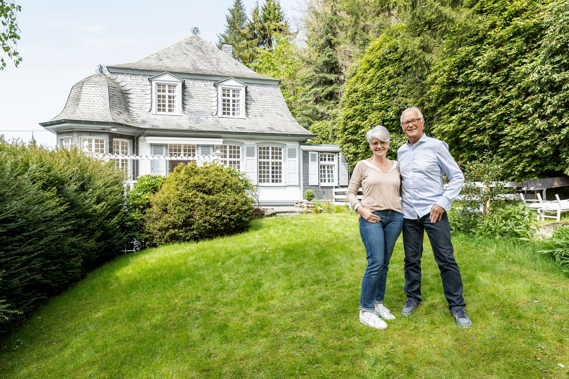 Couple standing in front of a house on a grassy hill, smiling. Green trees and a cloudy sky in the background.