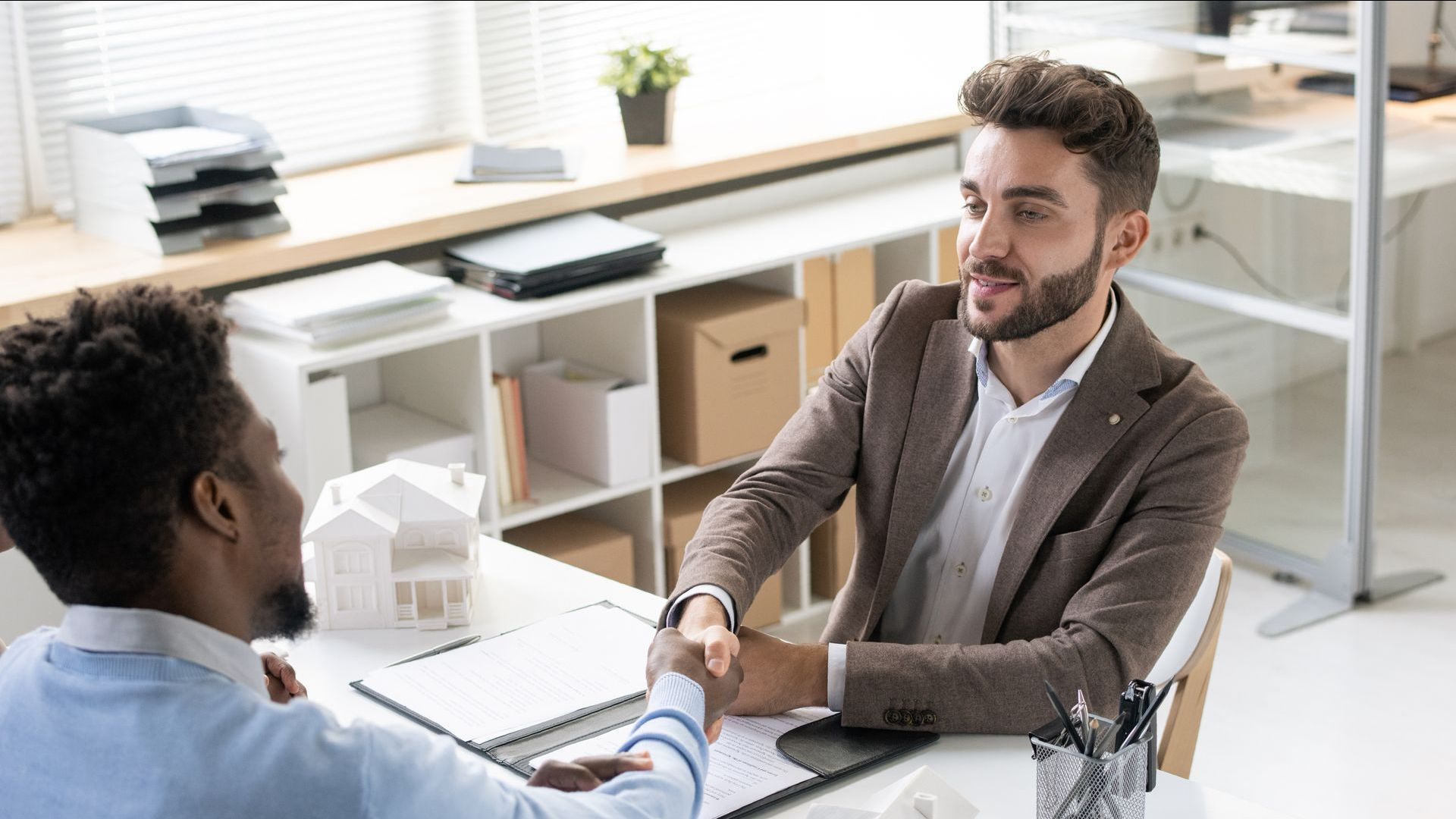 Two men shaking hands at a desk, possibly closing a deal. White office setting.