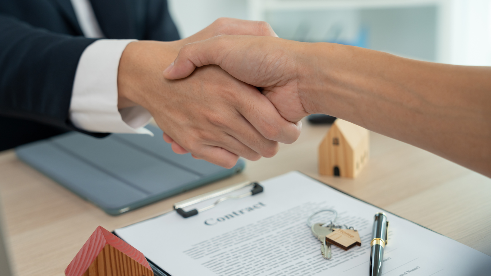 Two people shake hands over a contract with a small house model and a set of keys on the desk, symbolizing real estate.