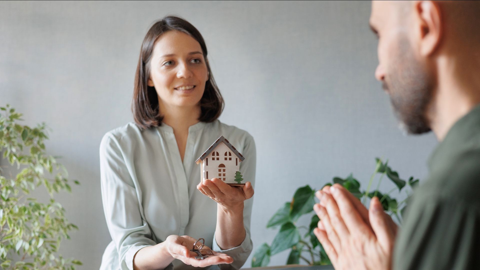 A woman holds a model house and keys while a man opposite her makes a thankful gesture.