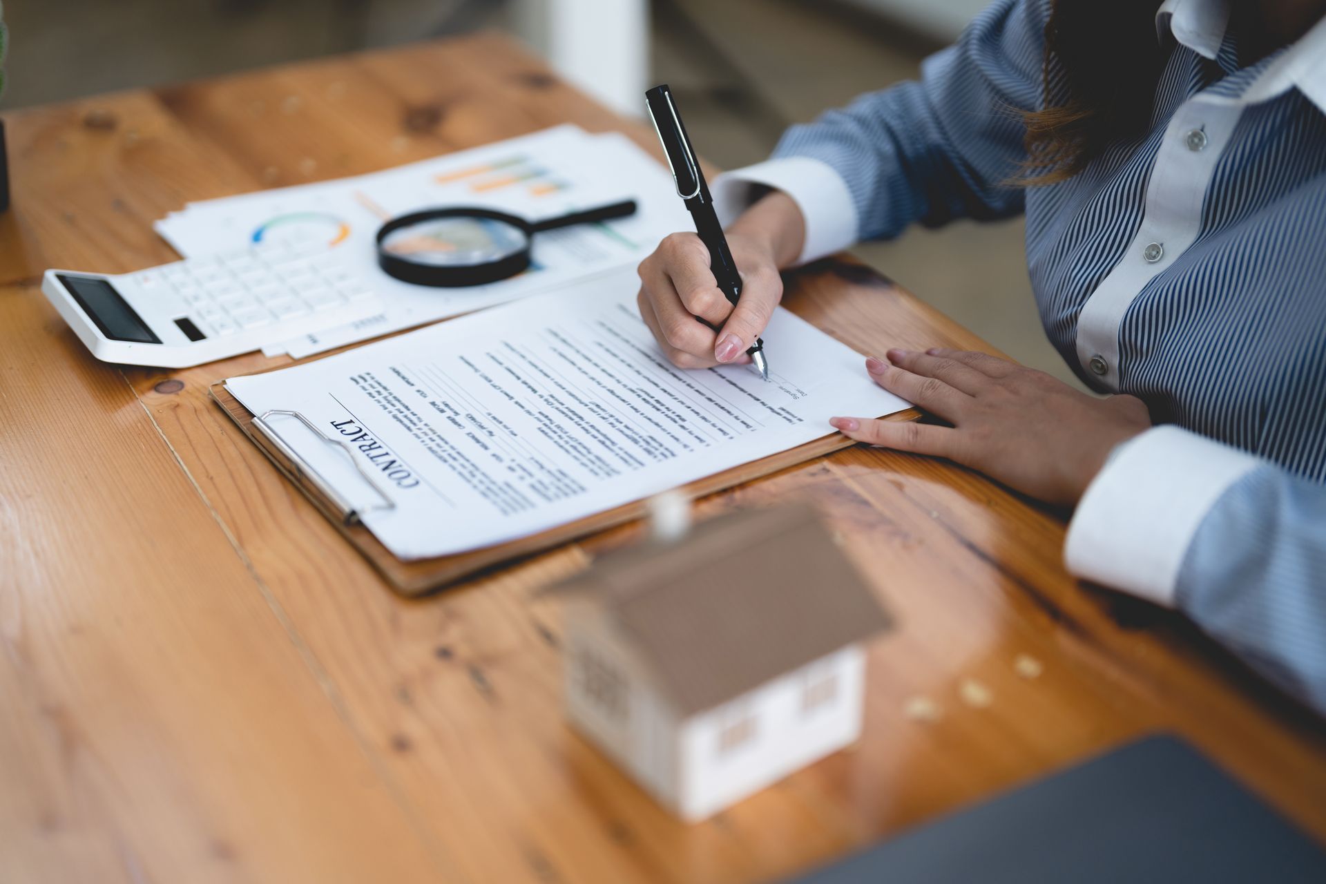 Woman signing documents on a clipboard, with a miniature house, calculator, and magnifying glass on a wooden desk.