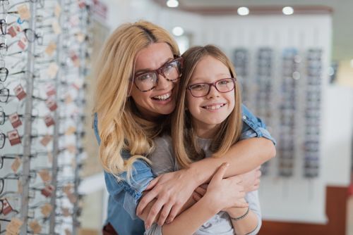 A woman and a girl wearing glasses are hugging each other in an optical shop.