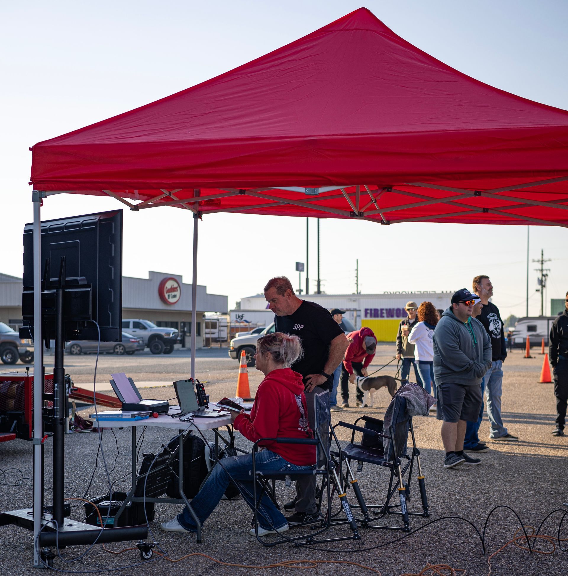 People under a red tent with monitors and equipment, watching an event outdoors.