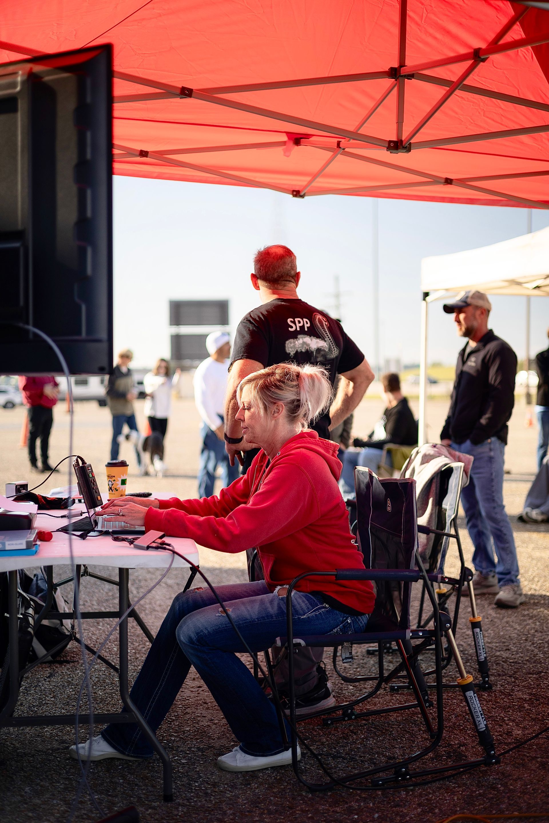Woman in red hoodie works on laptop under a red canopy. Other people stand nearby in an outdoor setting.
