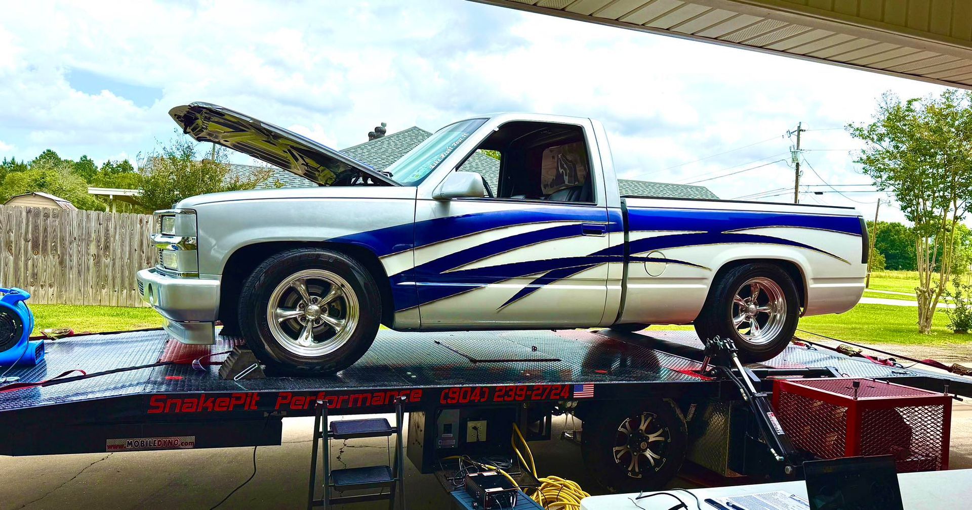 Silver pickup truck with blue flames, on a tow truck. Hood open, under a cloudy sky.