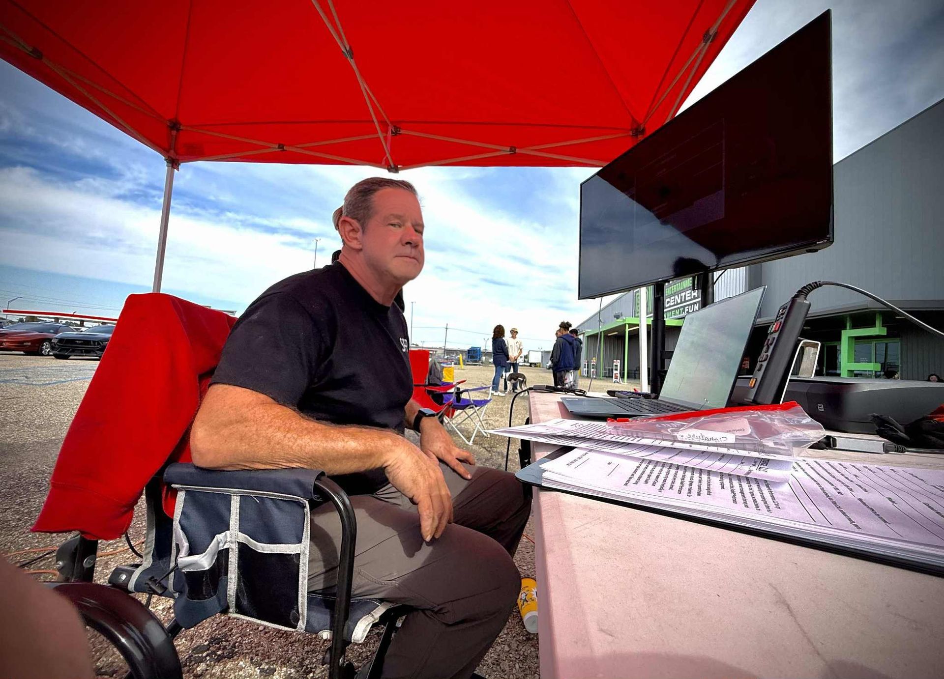 Man sitting under red canopy, looking at a laptop and large screen; outdoor setting.