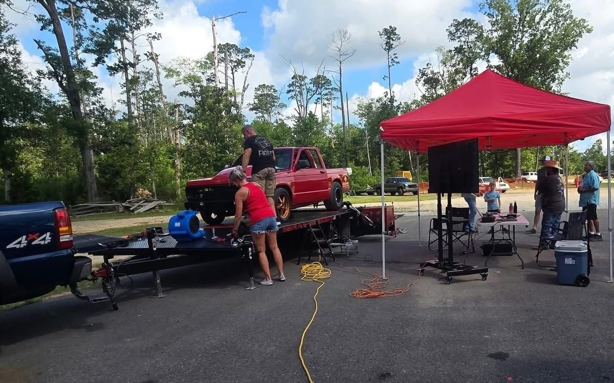 Red truck on a trailer at an outdoor event, with people and a red canopy.