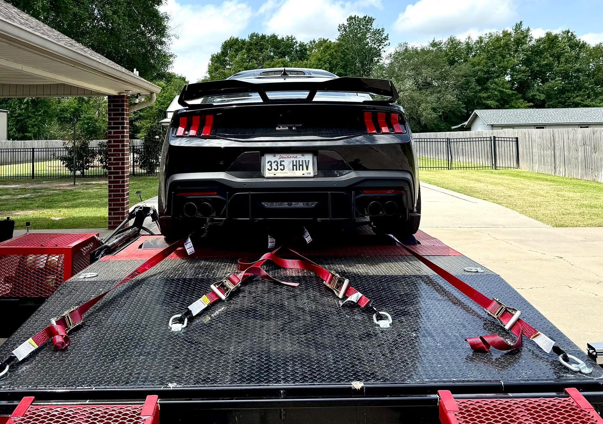 Black Mustang sports car secured on a black flatbed trailer with red straps, parked outdoors.
