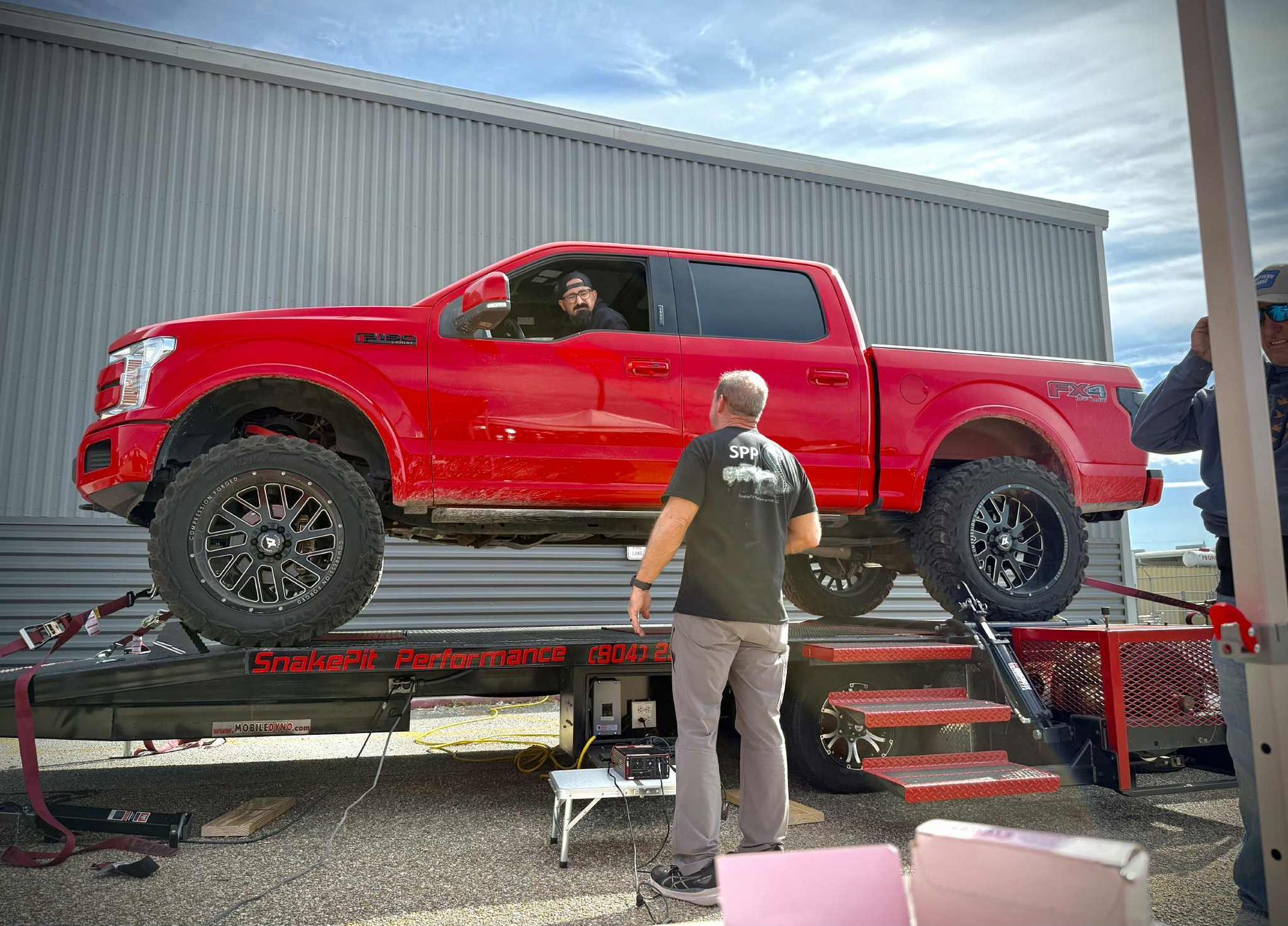 Red pickup truck on a lift; two men in front, one inside, against a metal building, outdoors.