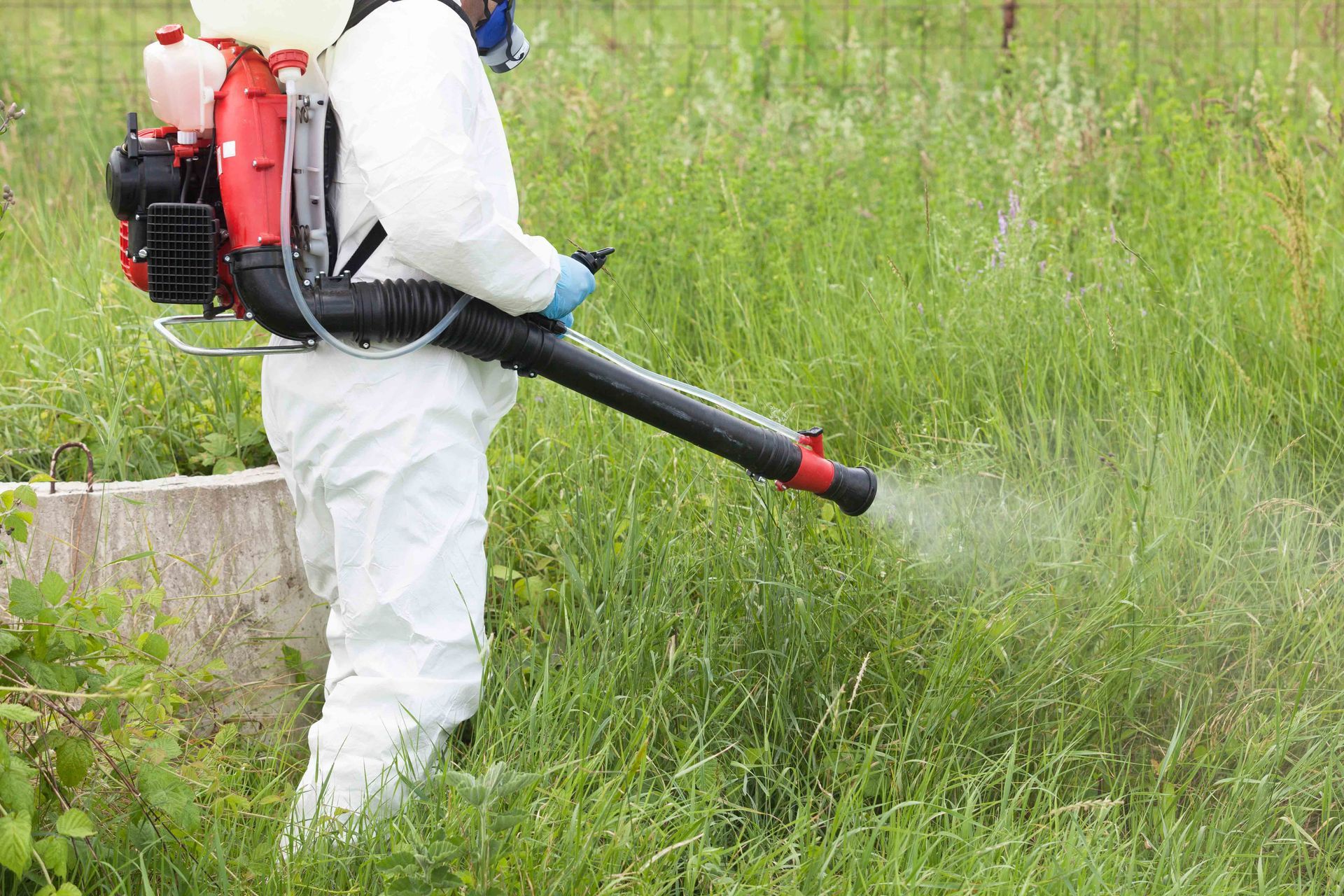 Photograph showing worker in white protective coveralls, gloves, and respirator using red backpack s