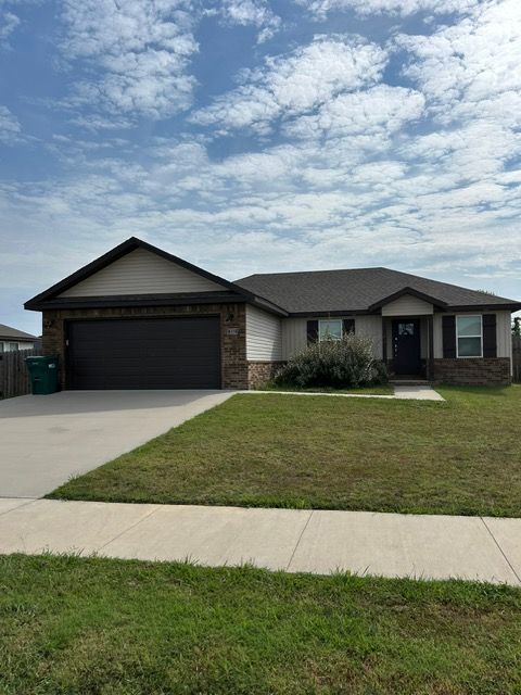 Tan and brown ranch-style house with a two-car garage, green lawn, and cloudy blue sky.