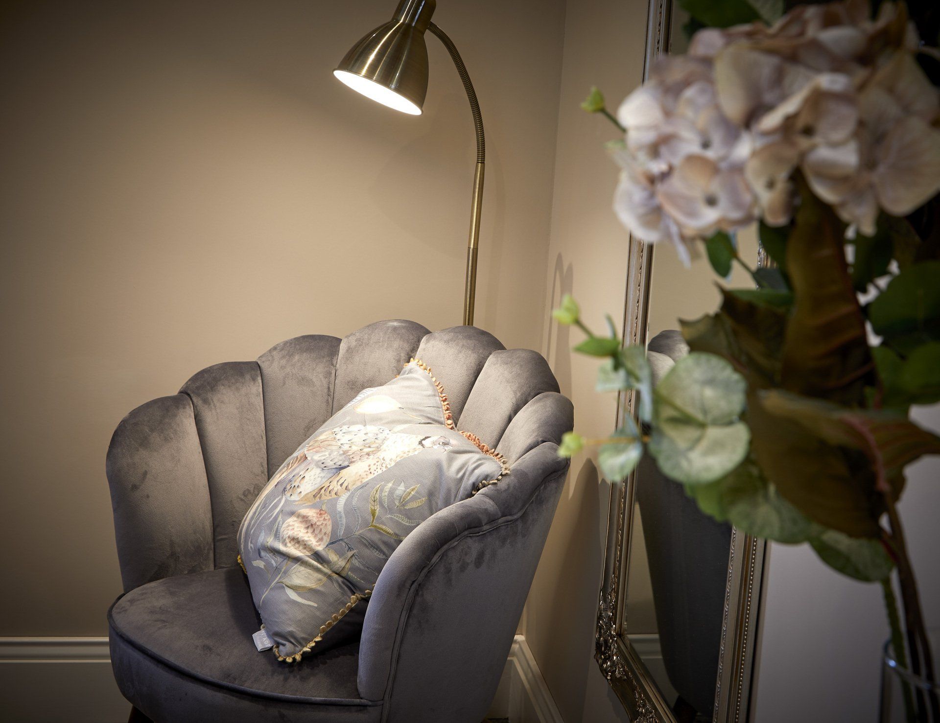 A chair with a pillow and a lamp next to a mirror and a vase of flowers.