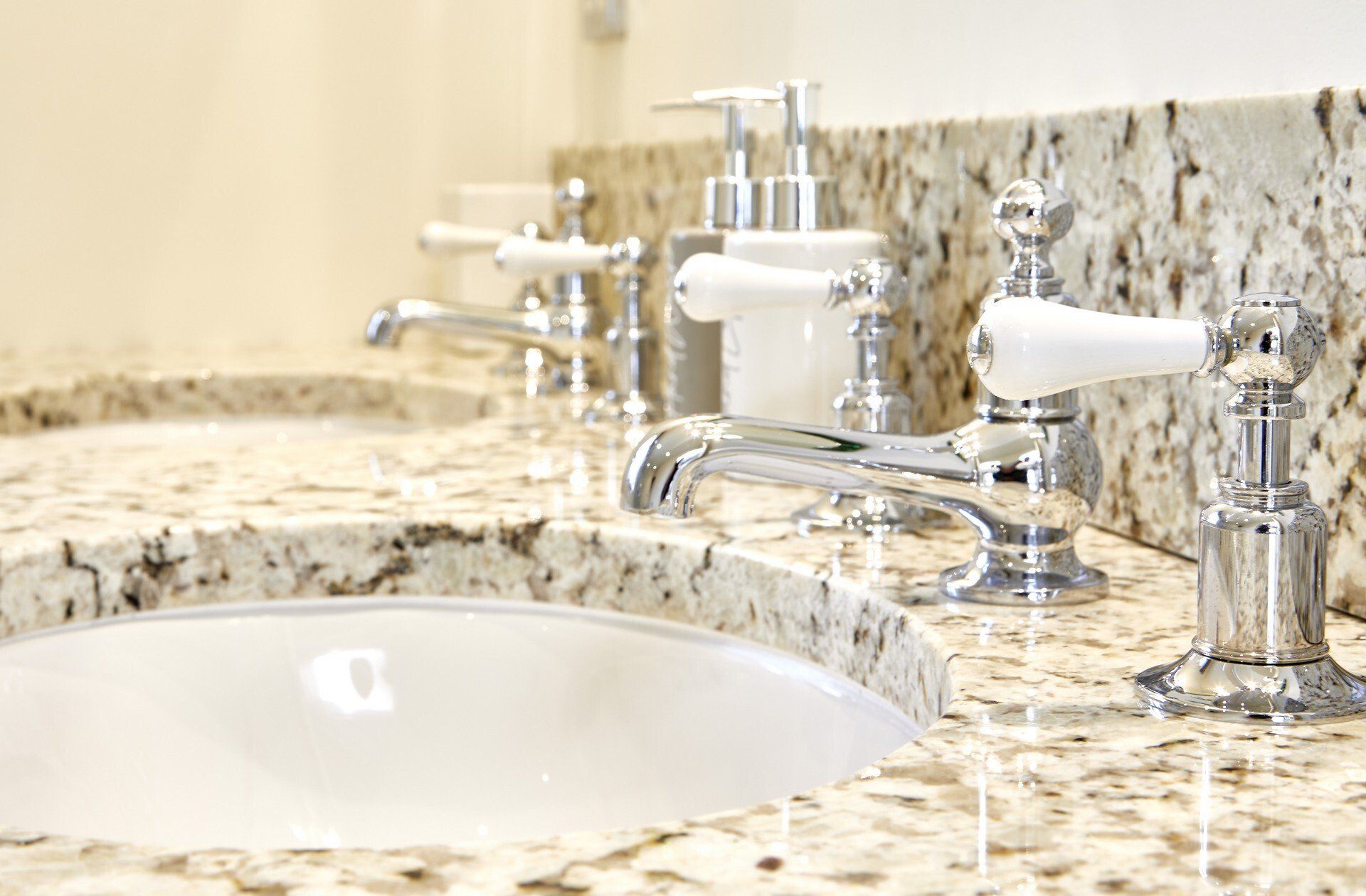 A bathroom sink with two faucets and soap dispensers on a granite counter top.