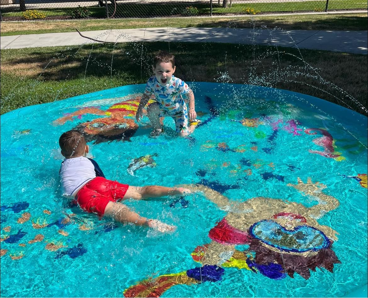 Two children are playing in a sprinkler pool in a park.