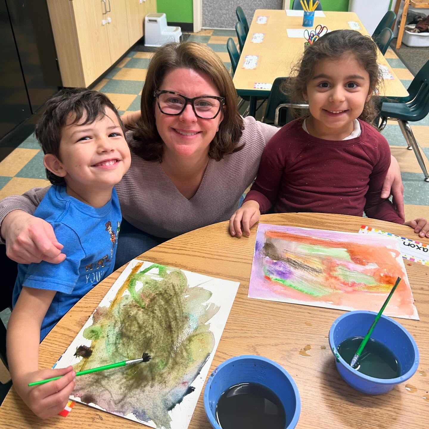 A woman and two children are sitting at a table with paintings and bowls of paint.
