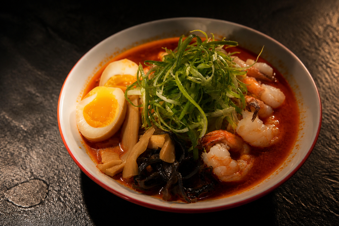 Red bowl of ramen with egg, shrimp, noodles, and greens on a dark table