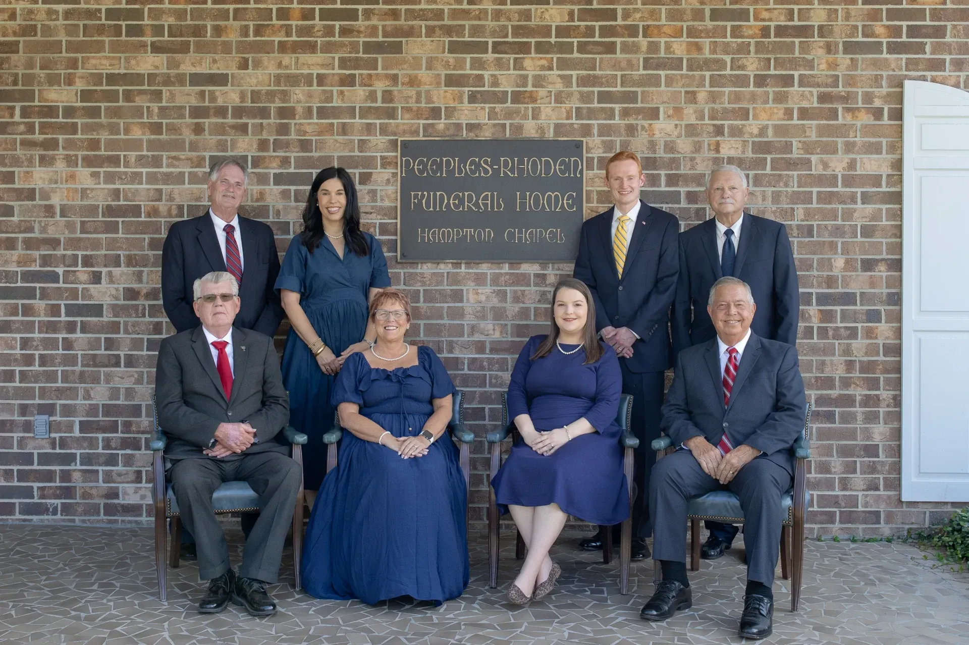 Group portrait of eight people posed outside a brick building, with a chalkboard sign behind them