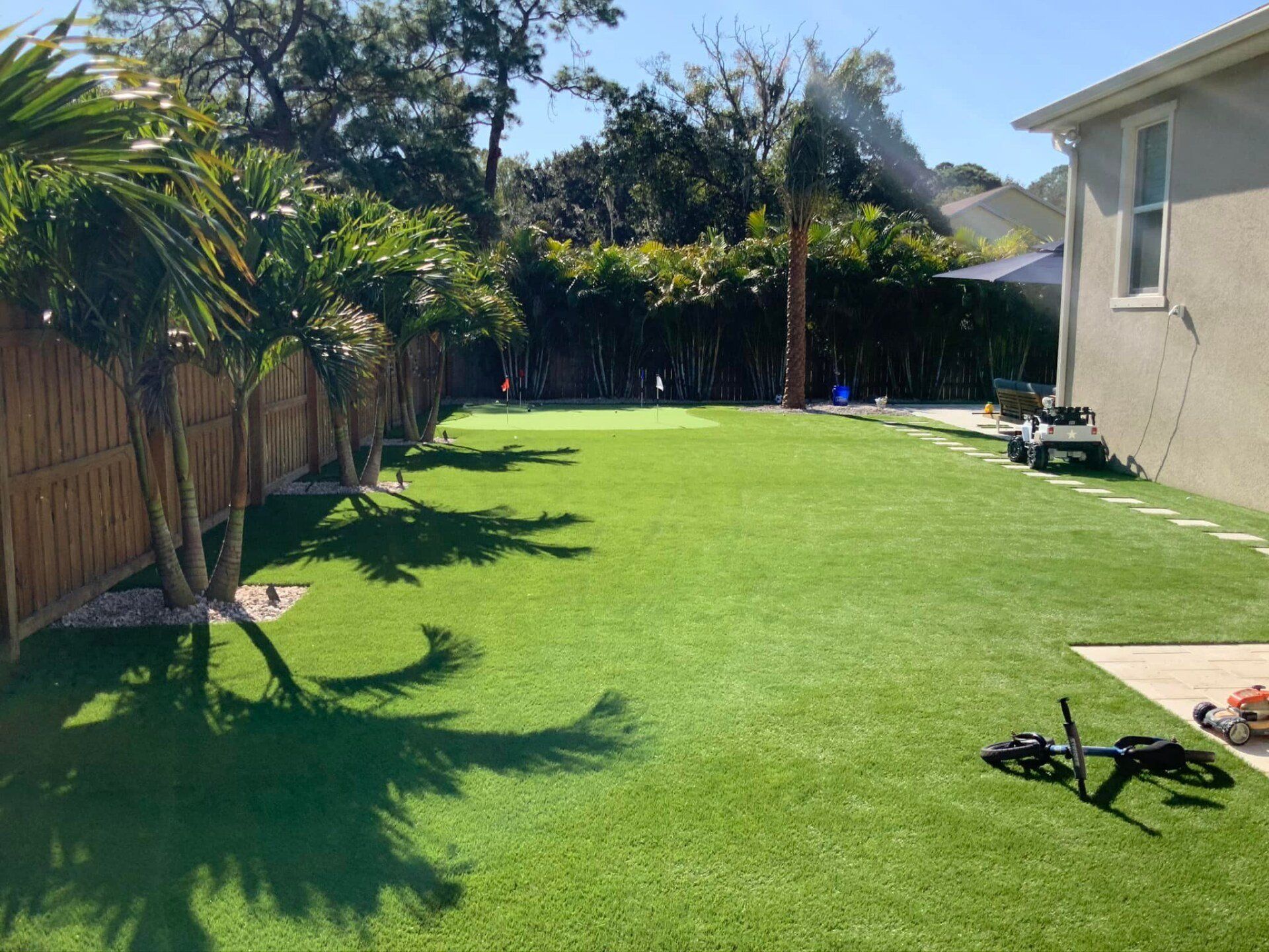 Green lawn with palm trees and a wooden fence. A lawnmower sits in the grass next to a house.