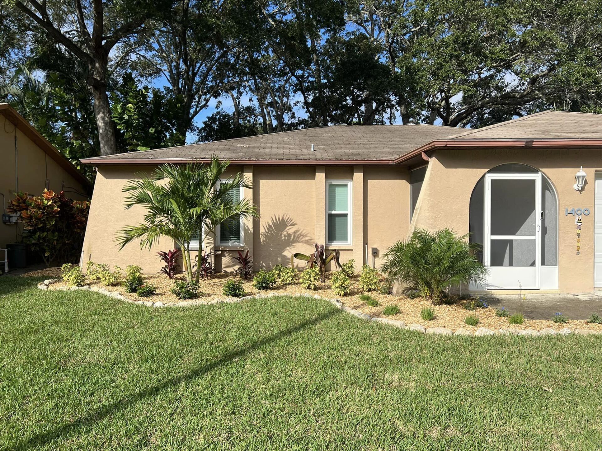Tan house with a green lawn and small garden beds in front. Palm trees and other greenery.